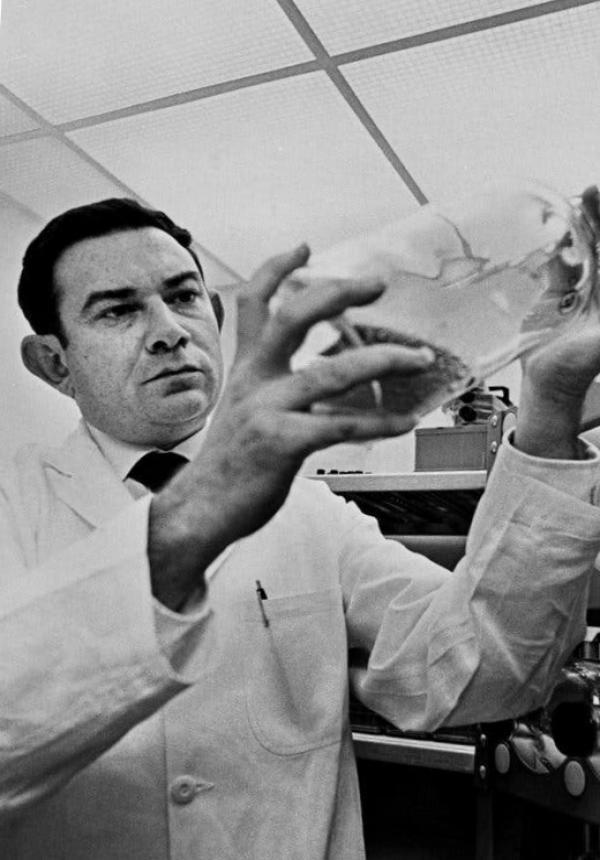 A black-and-white photo of Leonard Hayflick, a man in a white lab coat, holding up a glass container and looking at it intensely.