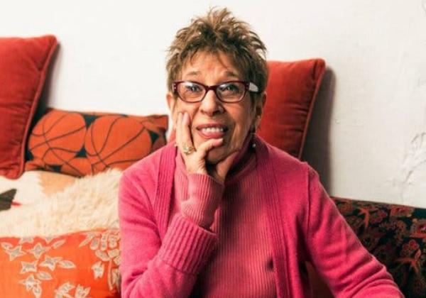 Hettie Jones, a woman wearing dark red eyeglasses and a pink sweater and resting her chin on her hand, sits on a couch with several red and orange pillows.