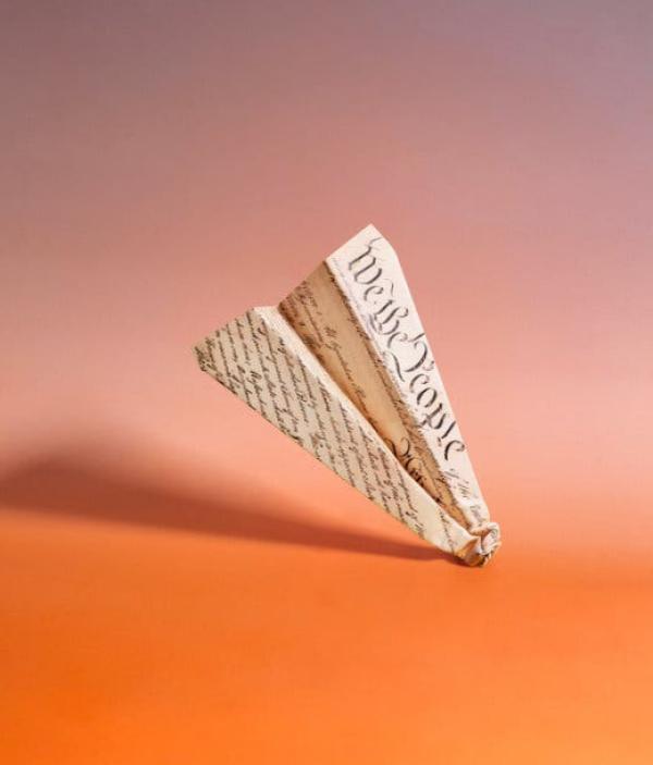 It is against a colorful background. Constitution crashing into the ground. A photo illustration shows a paper airplane made of the U.S.