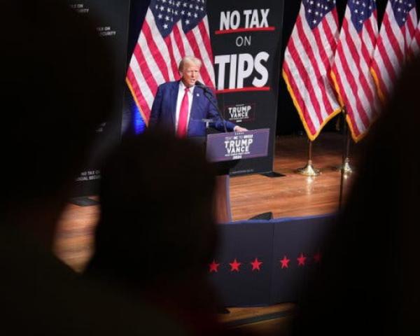 Donald Trump speaking on a stage in front of American flags.