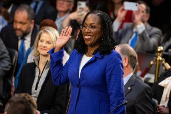 A 2022 photograph shows Ketanji Brown Jackson, a Black woman in a cobalt blue jacket, smiling broadly and lifting her right hand to take an oath at a congressional hearing.