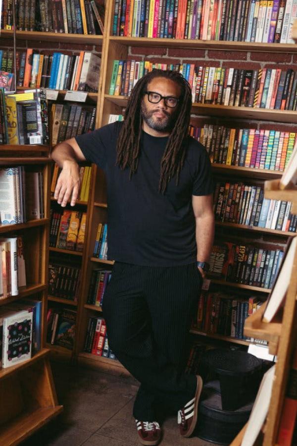 Franklin Leonard in a black shirt and pants poses for a portrait against bookshelves.