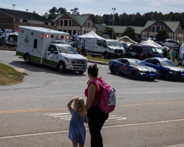 An ambulance and police cars are seen in front of school buildings with a child and woman looking at the scene from across a street.