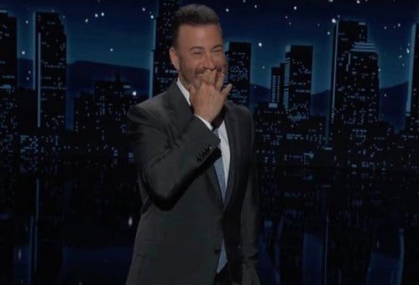 Jimmy Kimmel, standing against a stylized background of a nighttime skyline, laughs with a hand to his mouth.