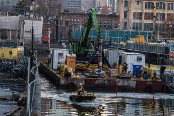 A worker on a small boat in the Gowanus Canal next to a barge, with buildings in the background.