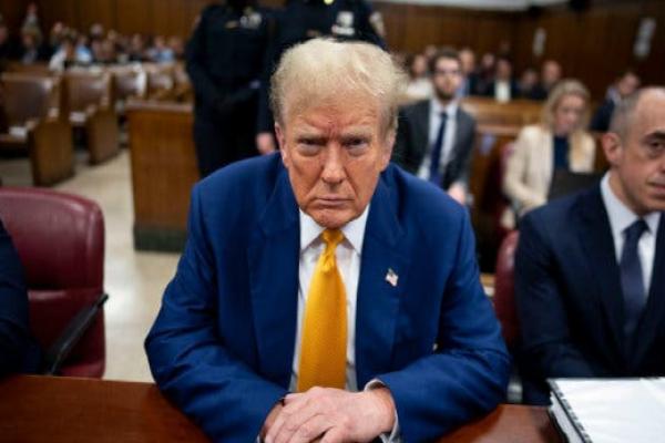 Trump sits at a table inside a courtroom and stares at the camera. Donald J.