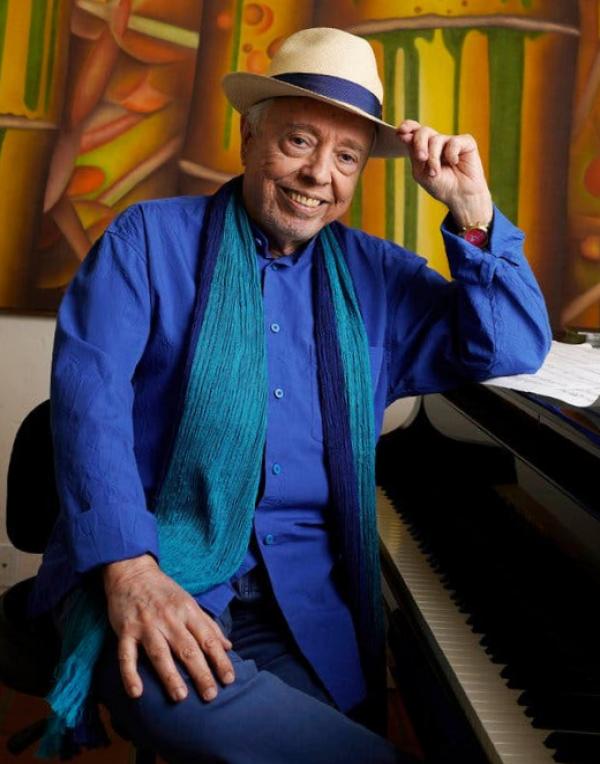 Sergio Mendes, dressed all in blue with a green scarf and a fedora, sits at a piano with his left elbow leaning on the top and smiles.