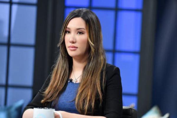 Lauren Chen sits a table, wearing a dark sweater, on what appears to be a TV set.