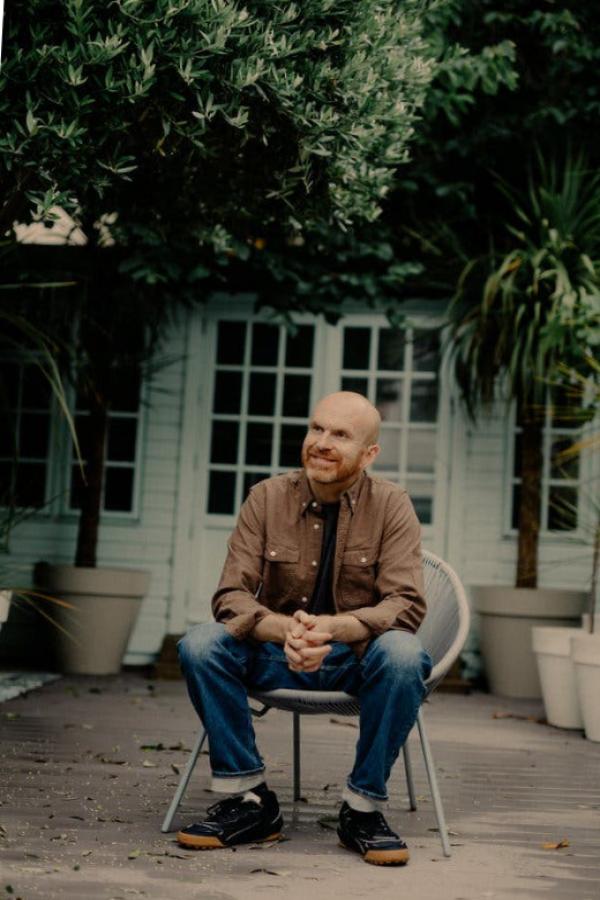 Haig sits on a chair, outdoors, with a white windowed doorway and shrubbery behind him.