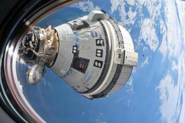 A white space capsule with the NASA logo floats outside a window with the cloud-streaked Earth behind it.