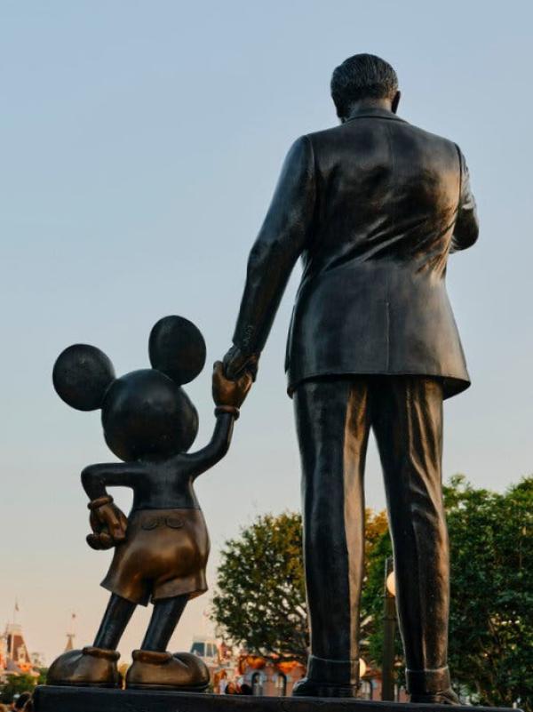 A statue of Mickey Mouse holding the hand of Walt Disney, seen from behind.