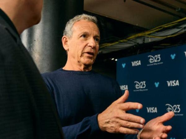 Bob Iger, wearing a blue sweater, gesturing while speaking in front of a backdrop with the Disney D23 and Visa logos on it.