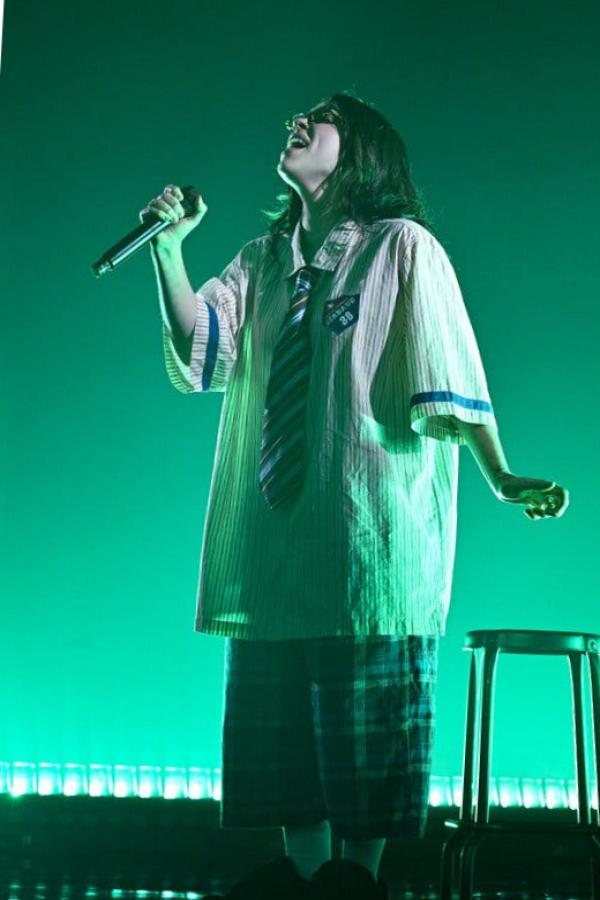 A woman in a baggy shirt and tie and long plaid shorts sings into a microphone onstage, bathed in greenish light.