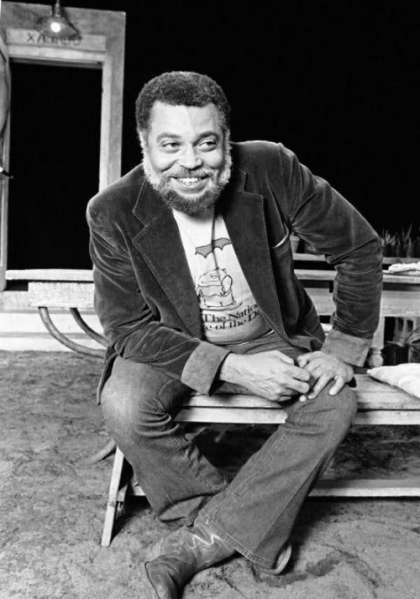 A black and white portrait of a young James Earl Jones sitting on a wooden bench and leaning forward and smiling with his hands on his left knee.