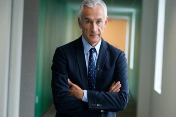 Jorge Ramos, wearing a tie and navy blue jacket, stands with his arms crossed in front of him.