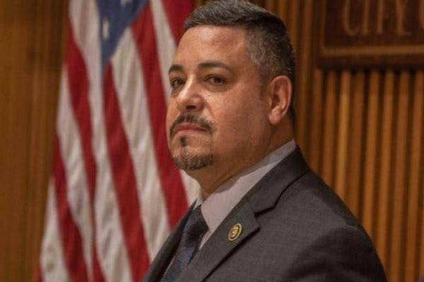 Edward Caban in a brown suit, behind Mayor Eric Adams who is speaking at a lectern.