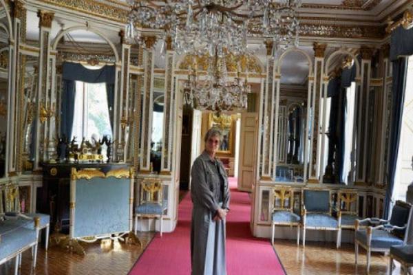 A woman wearing a gray outfit stands on a red carpet in an ornate room with chandeliers and gilded decorations.