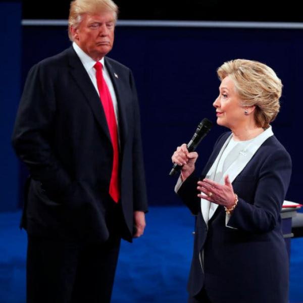 Hillary Clinton is standing and speaking into a microphone, as Donald Trump stands behind her looking on.