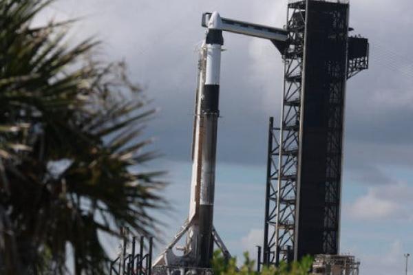 A SpaceX Falcon 9 rocket with the Crew Dragon capsule at the top standing at the launch stand in Florida on a partly cloudy day.