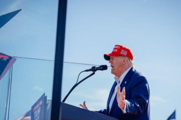 Trump, wearing a blue jacket, white shirt and red hat, stands at a lectern in front of a microphone. Former President Donald J.