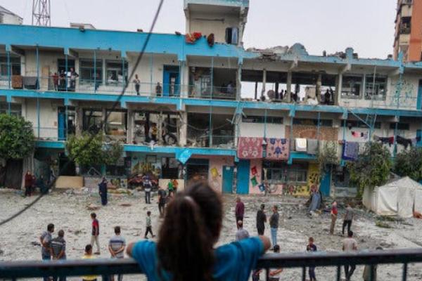 A girl watches people walking around the debris-strewn courtyard of a damaged school building.