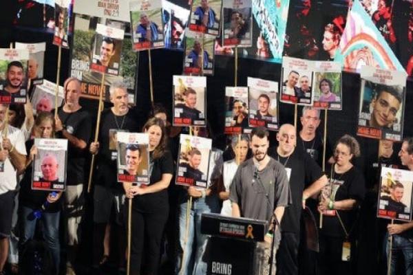 A crowd holding up posters bearing images of hostages being held in Gaza as a man speaks at a lectern bearing the words “Bring them home.”.