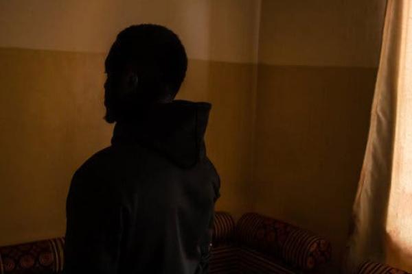 Jalil Muyeke stands inside a room, in the shadow, with orange and yellow walls behind him and a window.