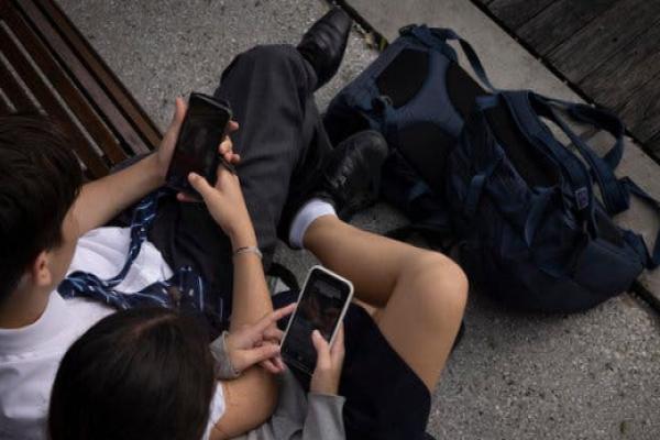 Two children in school uniforms, sitting on a bench and looking at their phones.