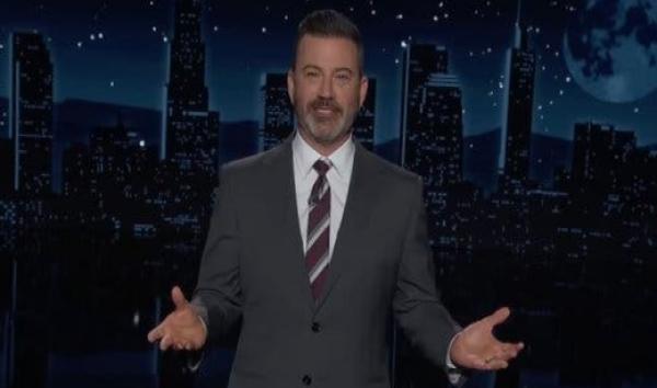 Jimmy Kimmel, wearing a gray suit, holds his hands out as he stands on the set of his show.