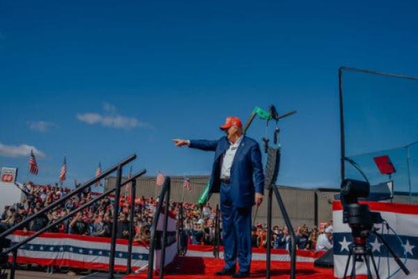 Trump points with a finger onstage at an outdoor rally, with crowd in the stands. Former President Donald J.