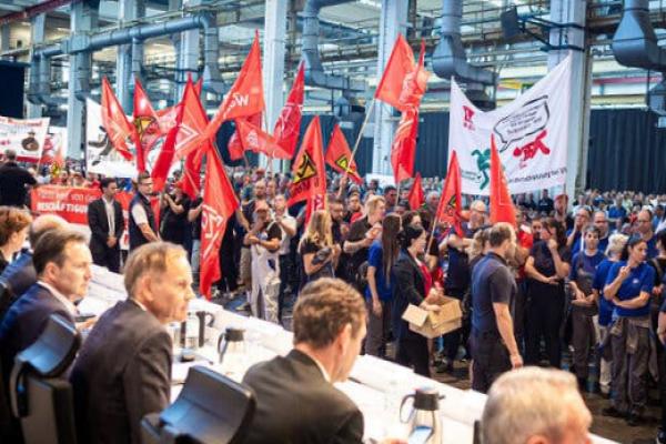 People with flags, inside a factory protesting in front of people seated behind a table.