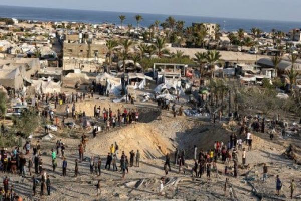 People cluster around two large craters in a sandy area surrounded by tents and low-rise buildings near the seaside.