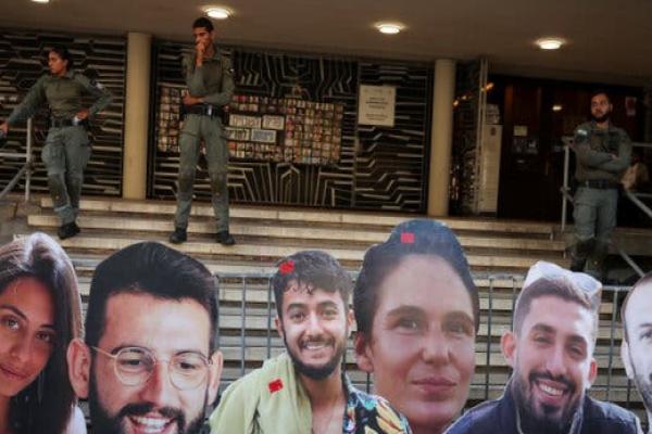 Enlarged photos of the six hostages recently found dead in Gaza stand in front of a barricade, with uniformed law enforcement personnel standing behind them.