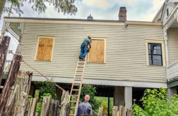 Another man watches from the ground. A man on a ladder boards up windows with plywood.