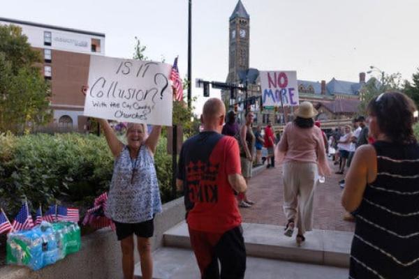 A woman dressed in shorts and a blue top holds a sign as several people walk by.