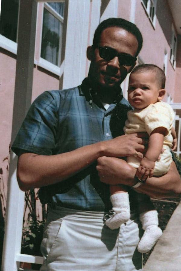 He wears a blue checkered short-sleeved shirt and sunglasses. Donald Harris holds a young Kamala Harris outside a pink stucco house.