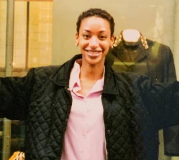 A woman wearing a pink shirt and a black coat stands in front of a window display, her arms outstretched.